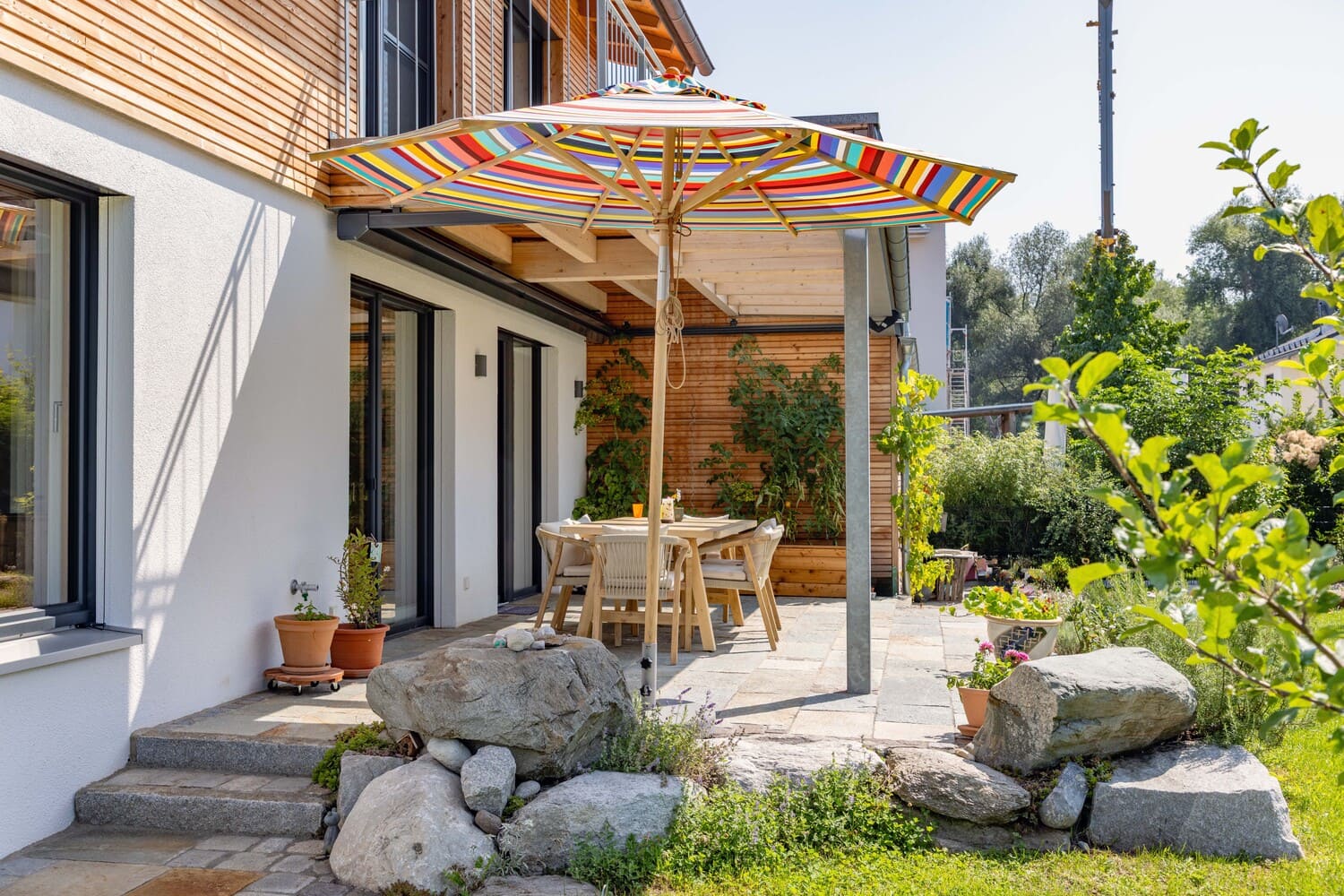 Eine sonnige Terrasse mit buntem Sonnenschirm, Holztisch und Stühlen lädt zum Entspannen im Garten ein.