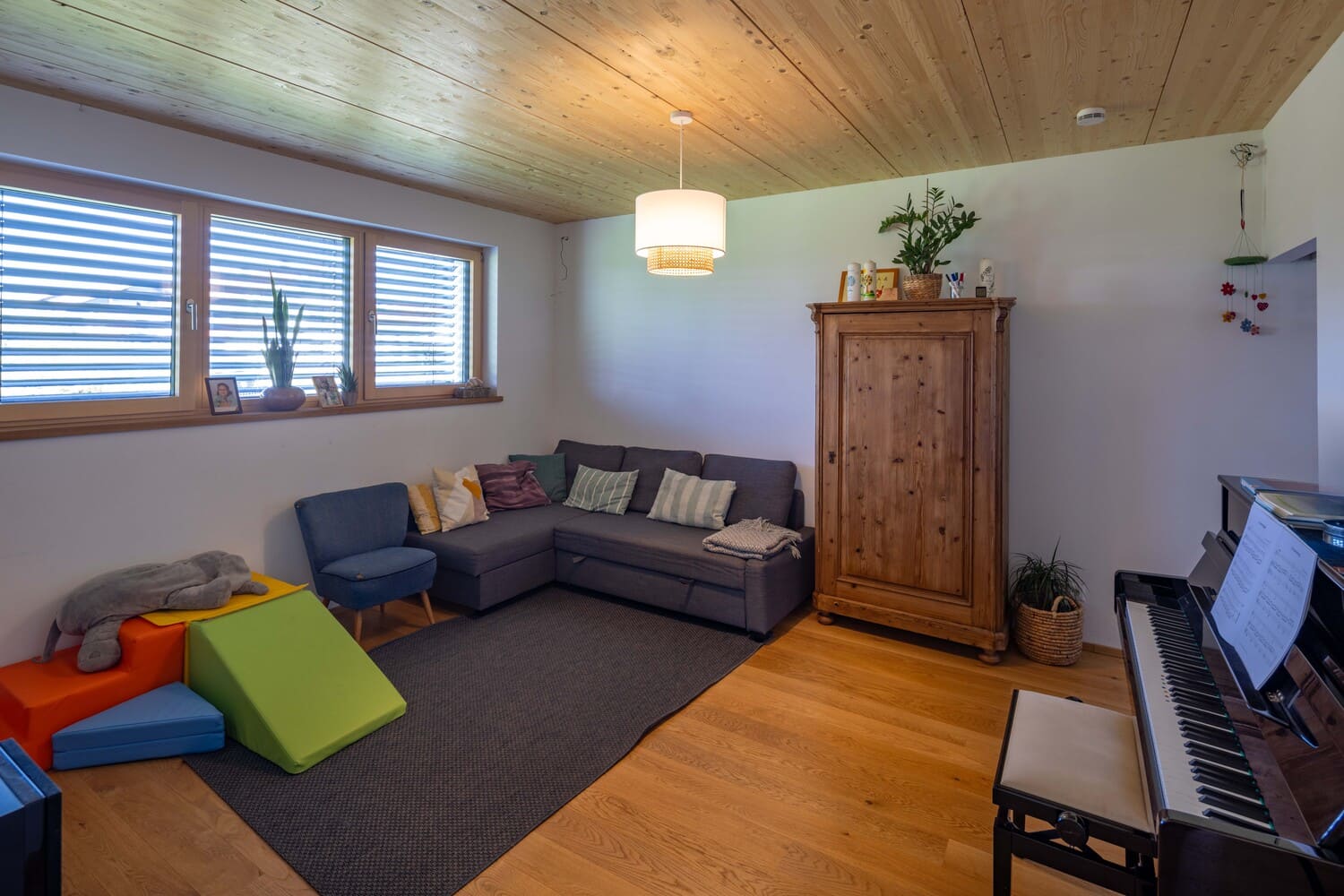 Ein gemütliches Wohnzimmer mit einem grauen Sofa, Holzmöbeln, bunten Spielmatten und einem Klavier lädt zum Entspannen ein.
