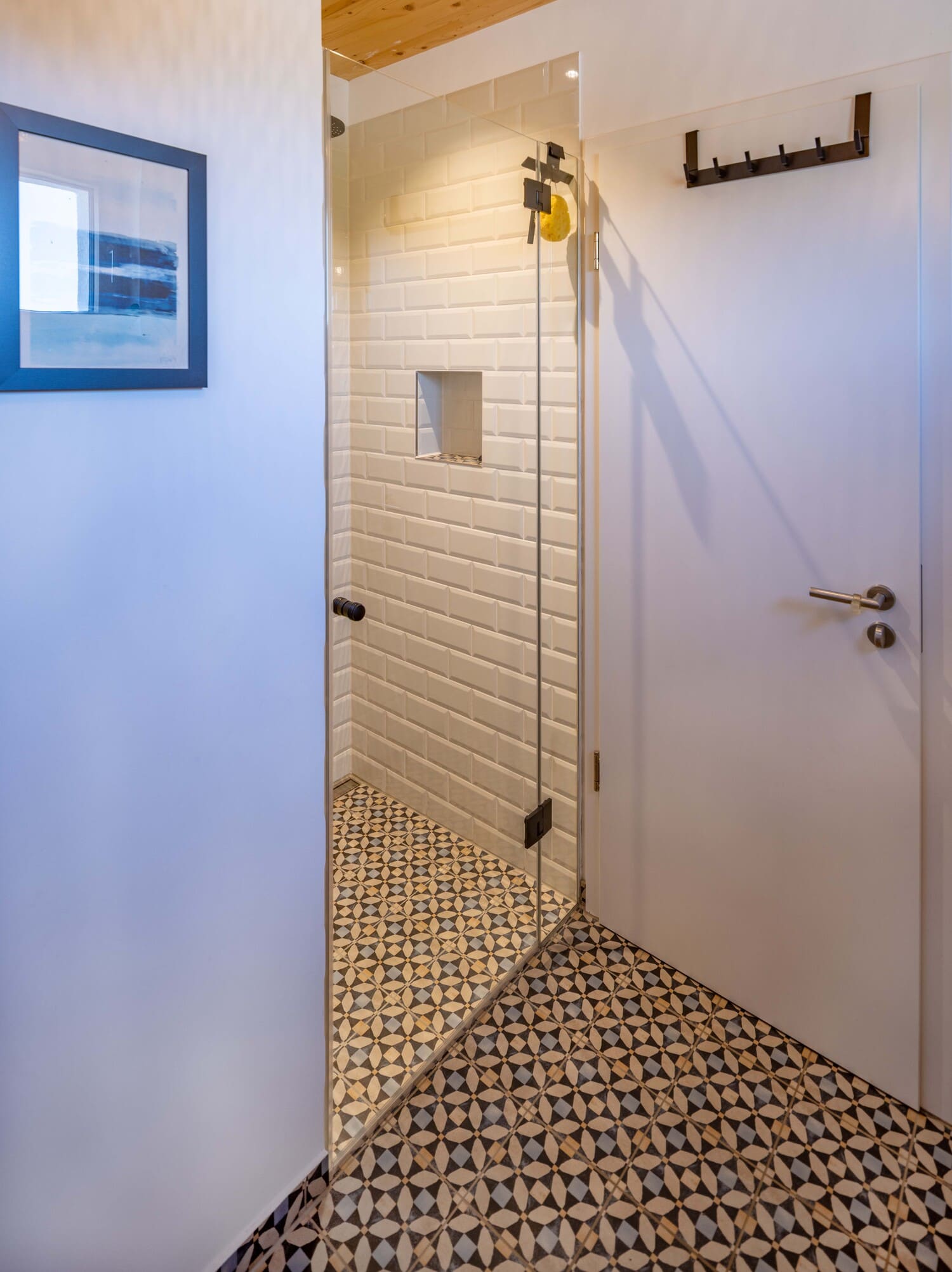 Eine moderne Dusche mit Glastür und weißen Fliesen steht auf einem gemusterten Boden neben einer geschlossenen Tür.