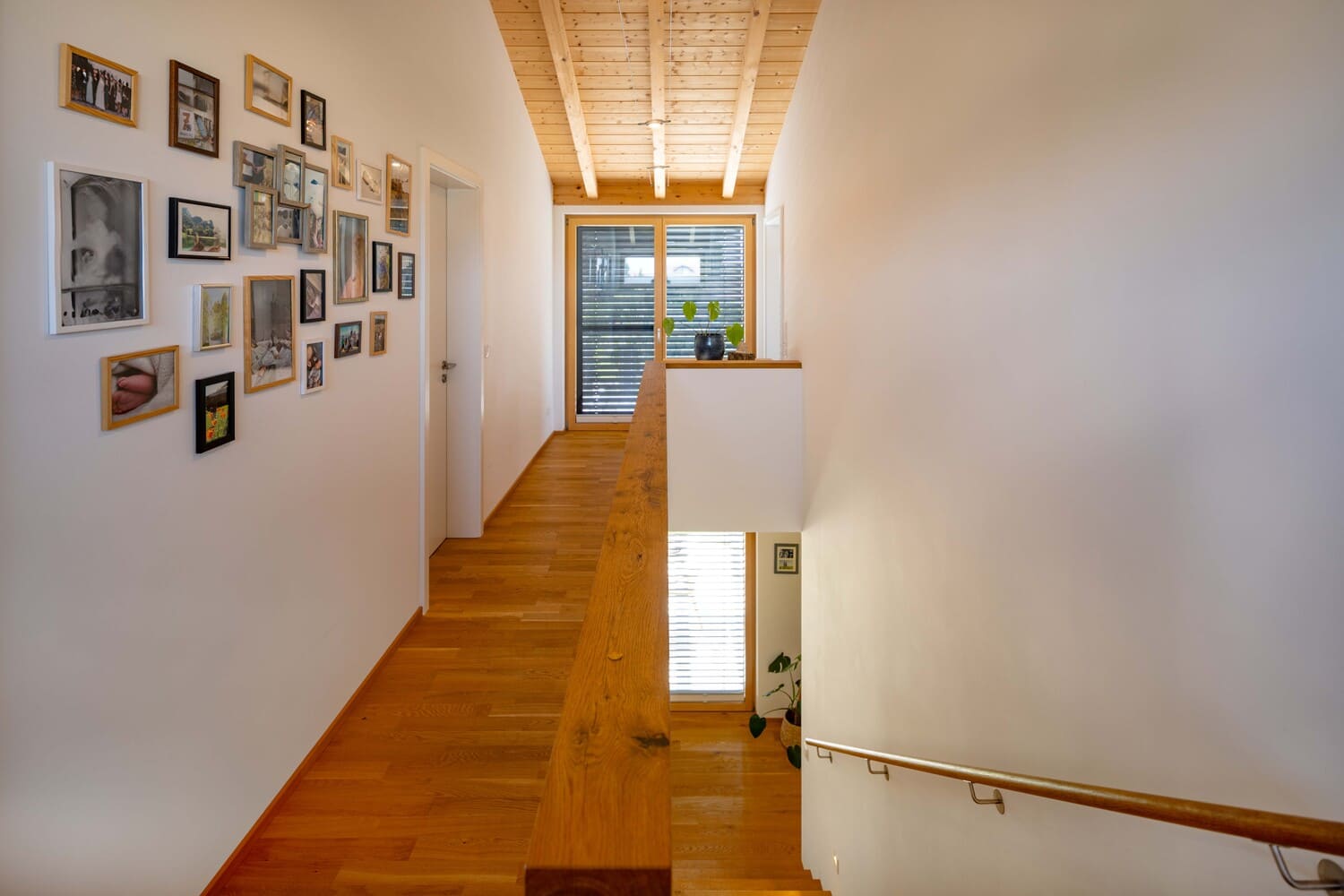Ein heller Flur mit Holzboden zeigt links eine Fotowand und am Ende ein großes Fenster, das Tageslicht hereinlässt.