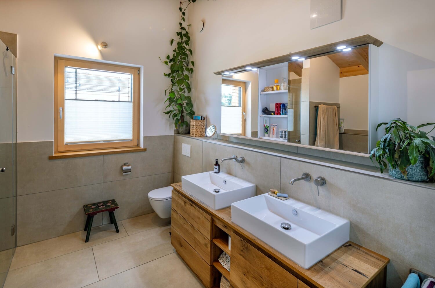 Ein modernes Badezimmer mit zwei Waschbecken, großen Spiegeln und üppigen grünen Pflanzen auf einer stilvollen Holzablage.